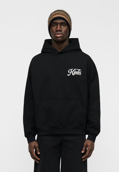 Μαύρο φούτερ με κουκούλα και τσέπη μπροστά, με λευκό λογότυπο στο στήθος, συνδυασμένο με σκούρο τζιν παντελόνι και ριγέ πλεκτό σκουφάκι.