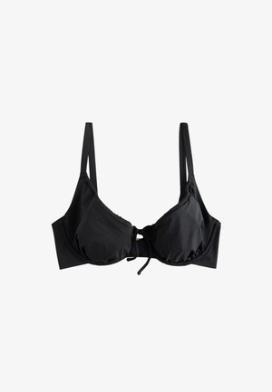 Mujer con el pelo mojado y un top de bikini negro está de pie en una playa de arena con el océano de fondo.
