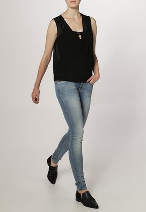 Femme marchant, portant un haut sans manches noir, un jean skinny bleu clair, des chaussures pointues noires et un collier pendentif en or.