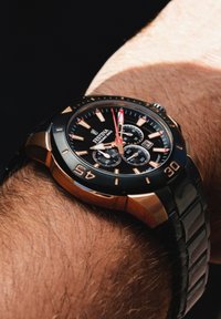 Orologio cronografo nero con dettagli dorati, grandi indicatori numerici e cinturino in gomma, indossato su un polso con pelle di un marrone chiaro.