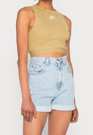 Kobieta ubrana w beżowy bezrękawnik Nike i jasnoniebieskie wysokie, podwinięte szorty jeansowe, stojąca na jasnym tle.