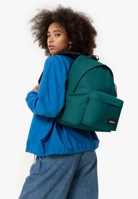 Mochila de tela verde con solapa superior, bolsillo frontal y parche con logo, llevada por una persona con chaqueta azul frente a un fondo blanco liso.