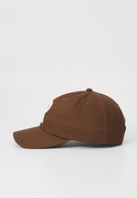 Brown cap with a curved brim, featuring a large white logo on the front, made from a soft fabric with visible stitching and eyelet vents.