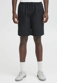 Shorts bleu marine avec une taille élastique et un cordon de serrage, présentant une coupe décontractée et un tissu lisse. Portés avec des baskets grises et des chaussettes blanches.