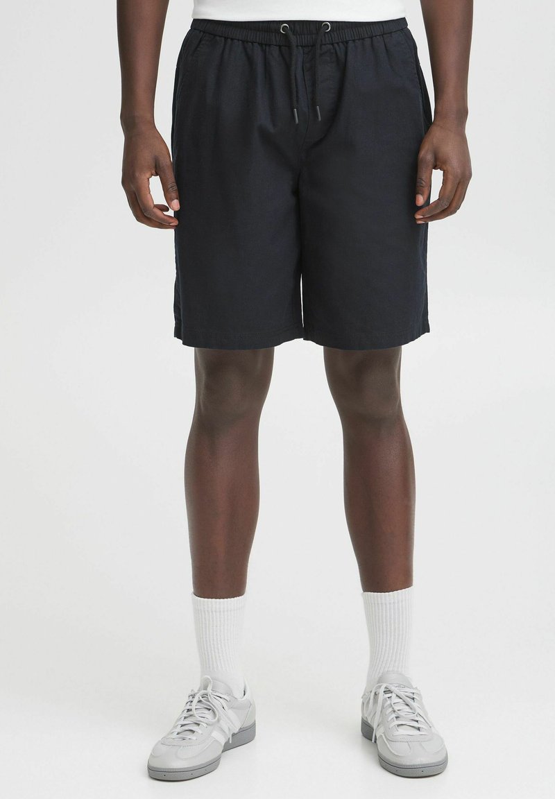 Shorts bleu marine avec une taille élastique et un cordon de serrage, présentant une coupe décontractée et un tissu lisse. Portés avec des baskets grises et des chaussettes blanches.