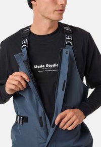 Wasserabweisende blaue Latzhose mit verstellbaren Trägern und Klettverschlussdetails, über einem schwarzen Langarmshirt mit weißen Schriftzügen.