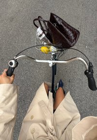Bicicletta con manubri cromati, un cestino in rete che tiene una borsa in pelle marrone a stampa coccodrillo e un limone, vista dall'alto, con un trench coat beige.