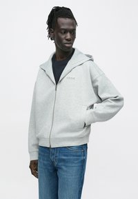 Γκρι φούτερ με φερμουάρ, από απαλό ύφασμα, με επενδεδυμένη κουκούλα, μπροστινές τσέπες και λεπτομέρεια λογοτύπου στο στήθος, συνδυασμένο με μπλε τζιν.