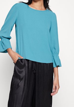 Frau trägt eine langärmelige türkisfarbene Bluse mit Rüschenmanschetten und lockere schwarze Satin-Hosen mit Kordelzug in der Taille.