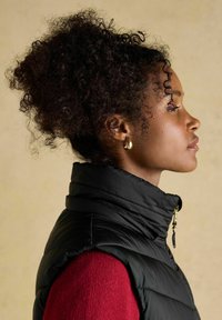 Czarna pikowana kamizelka z wysokim kołnierzem, z złotym zamkiem błyskawicznym. Noszona na czerwonym swetrze. Kręcone włosy ułożone do góry. Proste tło.