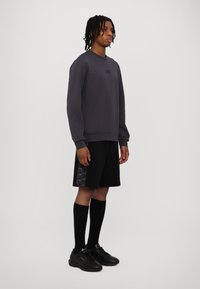 Felpa grigia con scollo rotondo, abbinata a pantaloni corti neri con logo stampato. Calzini neri e sneakers completano il look.