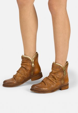 Bottes de cheville en cuir marron avec un design plissé, des détails en zip doré et un motif perforé sur la surface. Semelle plate.
