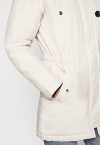Veste matelassée couleur crème avec taille élastique, poche latérale avec bouton-pression, et boutons-pression noirs sur la fermeture avant.