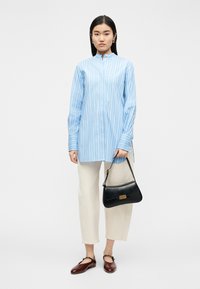 Γυναίκα με μπλε και λευκή ριγέ μπλούζα με μακριά μανίκια, κρεμ παντελόνι μέχρι τον αστράγαλο, καφέ παπούτσια Mary Jane και κρατώντας μια μαύρη τσάντα χειρός.