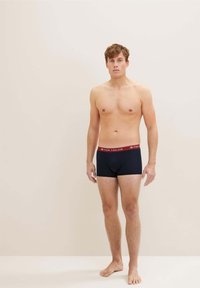 Črne boxer kratke hlače z rdečim elastičnim pasom, označenim "TOM TAILOR." Model stoji bos proti nevtralni ozadju.