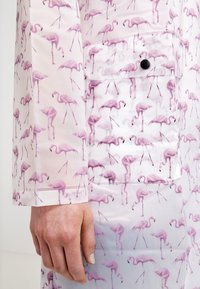 Regnrock med flamingomönster som har en blank finish, ljusrosa och vita färger, med en bröstficka och tryckknappsdetalj.