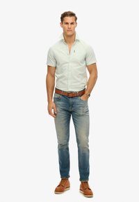 Camicia verde chiaro a maniche corte con tasca frontale, abbinata a jeans azzurri e stivaletti marroni con lacci, con una cintura e un orologio marroni.