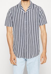 Muž v košili s krátkým rukávem, zapínací s vertikálními černobílými pruhy, oblečený v světle modrých džínech, stojící proti bílému pozadí.