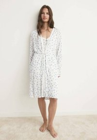 Robe florale blanche avec des motifs bleus, manches longues et ceinture à nouer. Texture lisse, longueur genou, portée par-dessus un top blanc.
