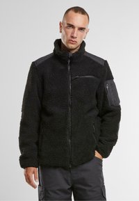 Giacca in pile nera con colletto alto, tasche con zip e dettagli a contrasto neri sulle spalle. Presenta una finitura testurizzata e pelosa.