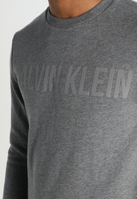 Sudadera gris con cuello redondo y mangas largas, con un destacado logo gris de Calvin Klein en el pecho.
