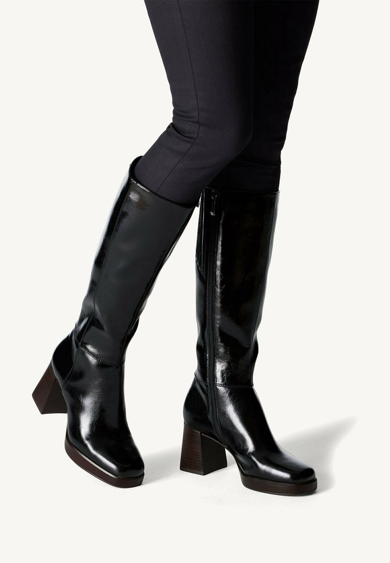 Shiny black knee-high leather boots with block heels worn with black fitted pants, shown against a plain light background.