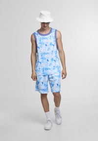 Mann som går, iført hvit bøttehatt, solbriller, blå og hvit marmorert tanktopp og matchende shorts, hvite sokker og joggesko.