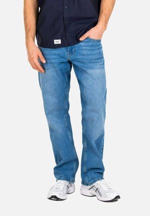 Man wearing light blue jeans, white sneakers, and a dark blue button-up shirt with one hand in pocket.