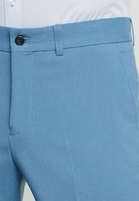 Pantalones azul claro con una textura suave, que cuentan con cierre de botón, trabillas para cinturón y bolsillos laterales. Corte ajustado con costuras visibles.