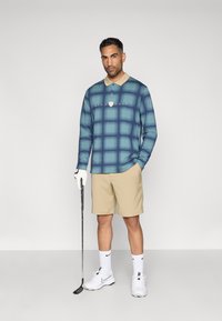 Polo de golf de manga larga a cuadros en azul con cuello beige, acompañado de pantalones cortos color crema. El modelo sostiene un palo de golf, vistiendo zapatos y calcetines blancos de Nike.