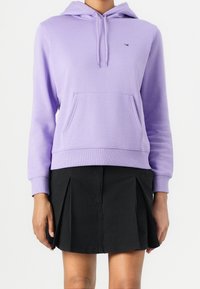 Sudadera lavanda con capucha, bolsillo canguro, puños y dobladillo acanalados. Combinada con una falda negra plisada. Diseño sencillo y casual.