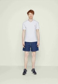 Man with curly red hair wearing a white short-sleeve V-neck shirt, blue denim shorts, and black sneakers standing against a plain background.