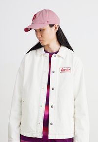 Jachetă din denim alb cu cusături roșii și închidere cu nasturi; prezintă un eticheta roșie "Dickies" pe pieptul stâng. Însoțită de o șapcă roz.