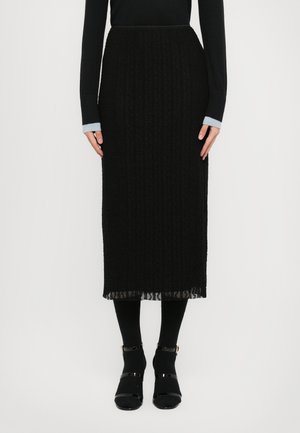 Femme portant une jupe midi noire texturée, un haut à manches longues noir avec des poignets gris, des collants noirs et des chaussures à bride de cheville noires.