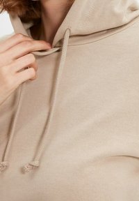 Sudadera beige con cordones, textura suave y un diseño simple. El material parece transpirable y tiene un corte relajado.