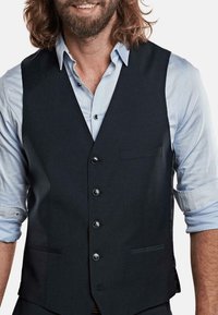 Schwarze Weste aus strukturiertem Stoff, mit sechs Knöpfen, einer Brusttasche und zwei Seitentaschen, getragen über einem hellblauen Hemd.