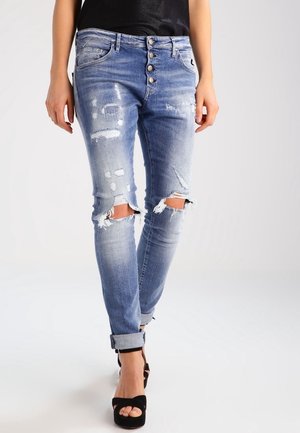 Kobieta ubrana w jasnoniebieskie, przetarte obcisłe dżinsy z rozdarciami na kolanach, podwinięte nogawki, czarną bluzkę i czarne sandały na wysokim obcasie idzie do przodu.