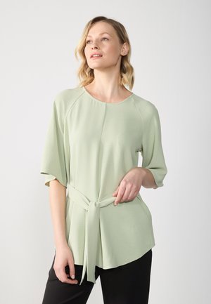 Top corto de color verde claro, con mangas cortas y tejido texturizado, con un escote redondo y un detalle de cintura atada. Combinado con pantalones negros.