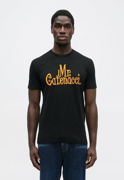 Černé tričko s krátkým rukávem, na kterém je velký oranžový nápis "Mr. Catenacci." Hladká látka s kulatým výstřihem, nošeno s modrými džíny.