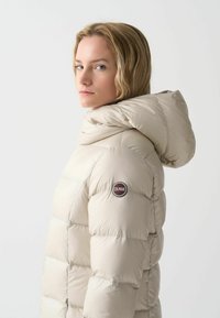 Bež jakna od pufastog materijala s visokim ovratnikom i pričvršćenom kapuljačom. Ima prošivene sekcije i kružni logo na lijevom rukavu. Glatka, sjajna tkanina.
