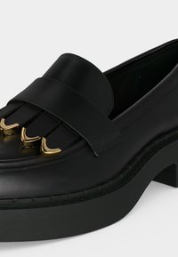 Mocassin noir en cuir avec semelle épaisse, accents décoratifs en métal doré sur le détail à franges avant.