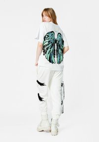 Camiseta blanca con gráfico de alas en negro y verde en la parte posterior, combinada con joggers blancos que tienen detalles en negro y botas blancas.