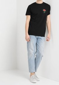 Osoba v černém tričku s vyšívanou červenou růží na hrudi, světlých modrých džínách a bílých teniskách, stojící proti bílým zdím.