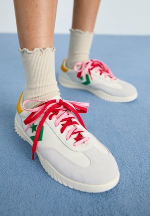 Legs wearing beige ruffled socks and white sneakers with green star logos and mismatched pink and red shoelaces on a blue floor.