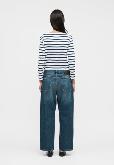 Ριγέ μπλούζα με μακριά μανίκια σε λευκό και ναυτικό μπλε, συνδυασμένη με τζιν παντελόνι φαρδύ στον ποδόγυρο σε σκούρο μπλε, με πίσω τσέπες και υφή.