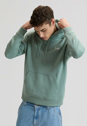 Grüner Kapuzenpullover aus Baumwolle mit Kängurutasche vorne, Kordelzug an der Kapuze und eingesticktem "Lee"-Logo. Modell trägt locker sitzende hellblaue Jeans.
