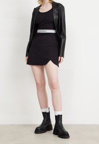 Nainen, jolla on päällään musta olkaimeton mini mekko, Tommy Jeans -vyö, musta nahkatakki, valkoiset sukat ja mustat paksupohjaiset saappaat.