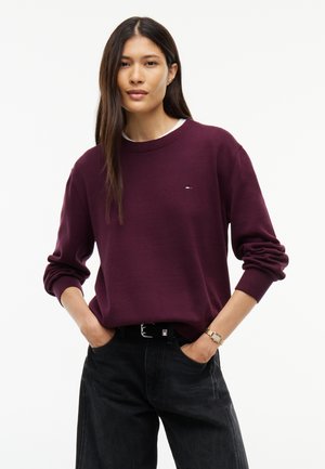 Frau mit langen dunklen Haaren, die einen bordeauxroten Pullover, schwarze Hose und eine goldene Uhr trägt, steht mit den Händen in den Taschen vor weißem Hintergrund.