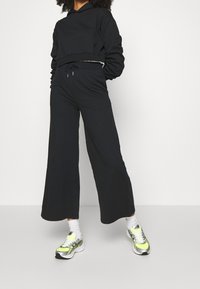 Svart kort hoodie med vida svarta byxor, som har elastisk midja och dragsko. Kombinerad med träningsskor med neondetaljer.