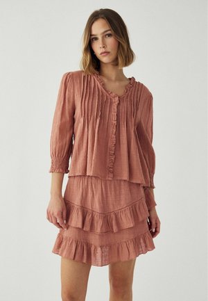 Žena oblečená v dvojdielnom outfite v prachovo ružovej farbe, pozostávajúcom z volánovej blúzky so zapínaním na gombíky a zodpovedajúcej vrstvenej sukne s volánmi, stojaca pred jednoduchým pozadím.